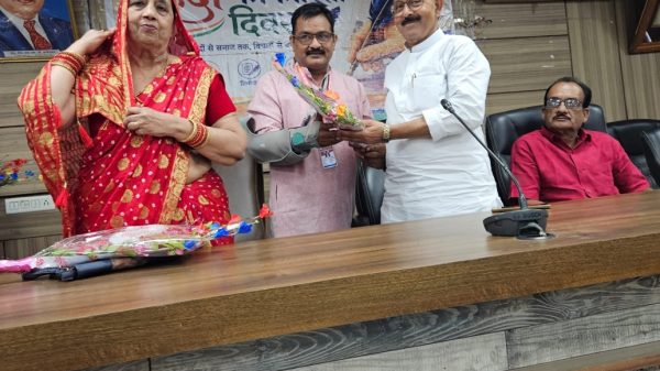Woman in a red sari presents a bouquet to a man in a white kurta at a formal event, with a banner and portraits in the background.