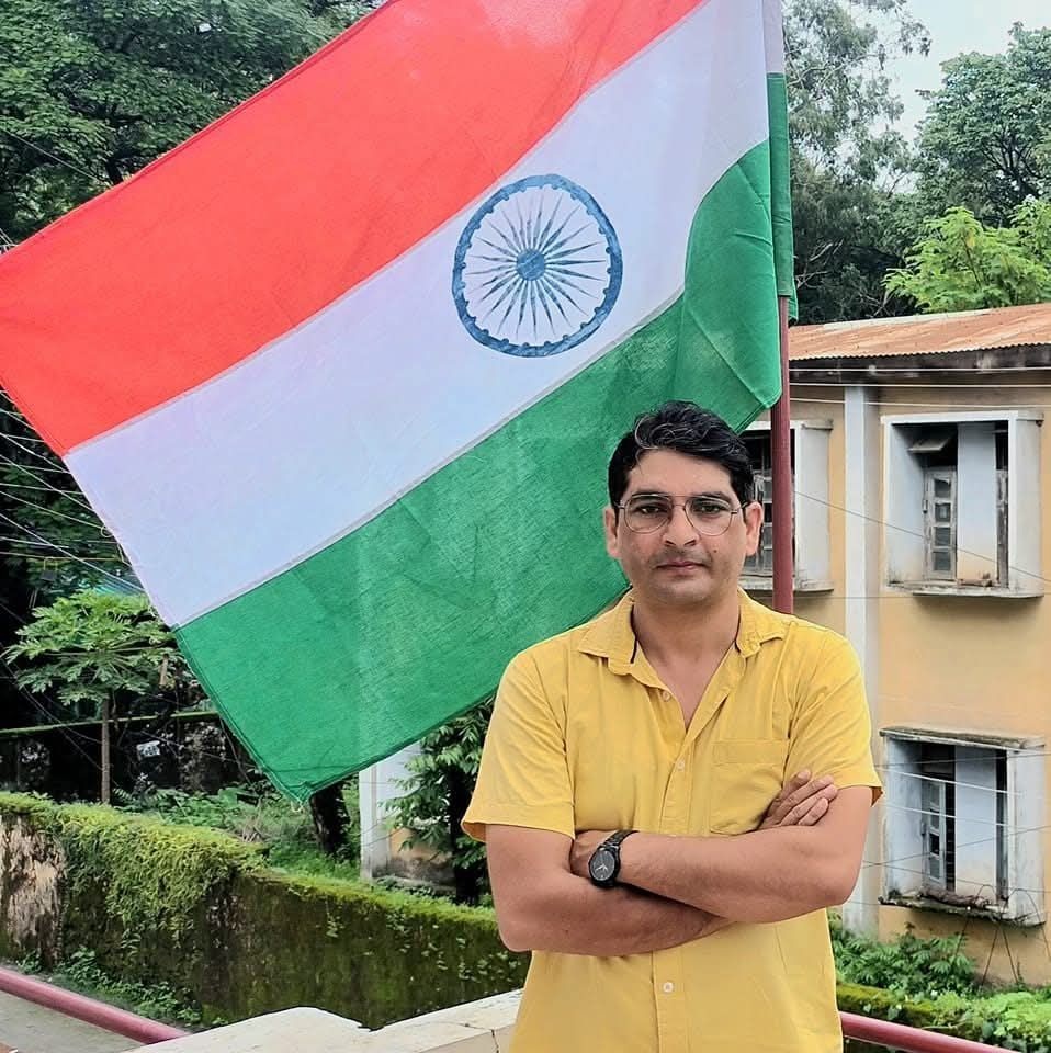 hem bhatt_ | No. 1 Indian Media News Portal Man in a yellow shirt stands with arms crossed on a balcony, waving Indian flag in the background.