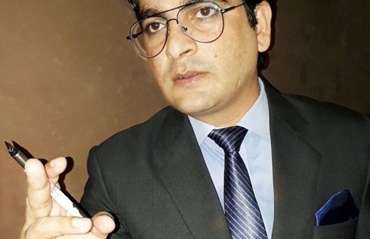 Man in a dark suit and glasses holding a marker and pointing toward the camera, as if presenting.