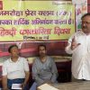 Three men in a room with a yellow banner in Hindi announcing Hindi Journalism Day (May 30); one man stands gesturing while two sit and listen.