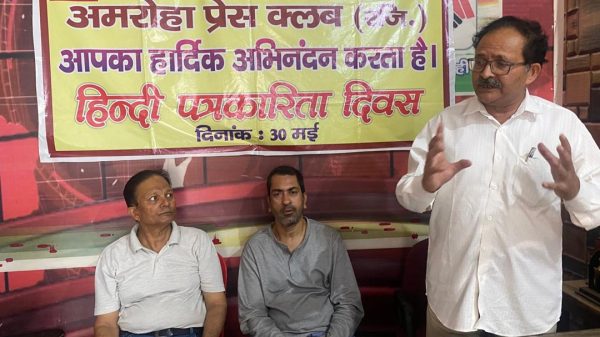 Three men in a room with a yellow banner in Hindi announcing Hindi Journalism Day (May 30); one man stands gesturing while two sit and listen.