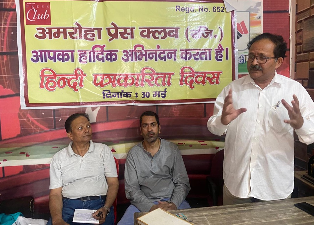 Three men in a room with a yellow banner in Hindi announcing Hindi Journalism Day (May 30); one man stands gesturing while two sit and listen.