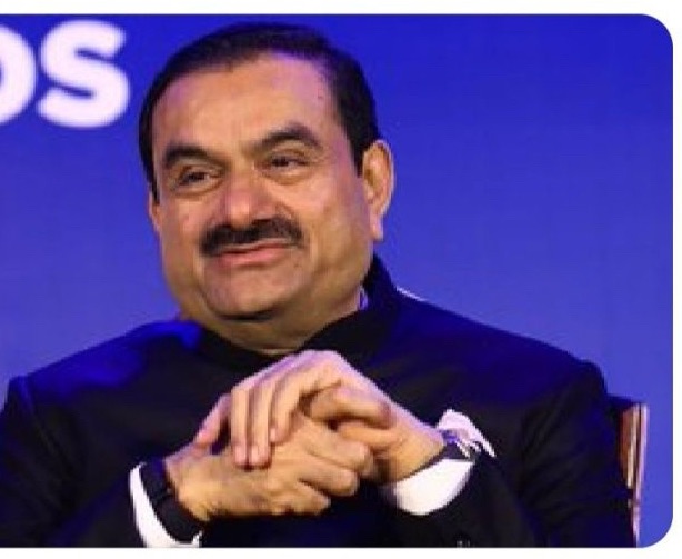 Smiling man in a dark suit sits with his hands clasped on a stage against a blue backdrop.