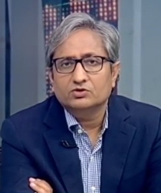 Male guest with gray hair and glasses, wearing a blue blazer and checkered shirt, speaking on a television set.