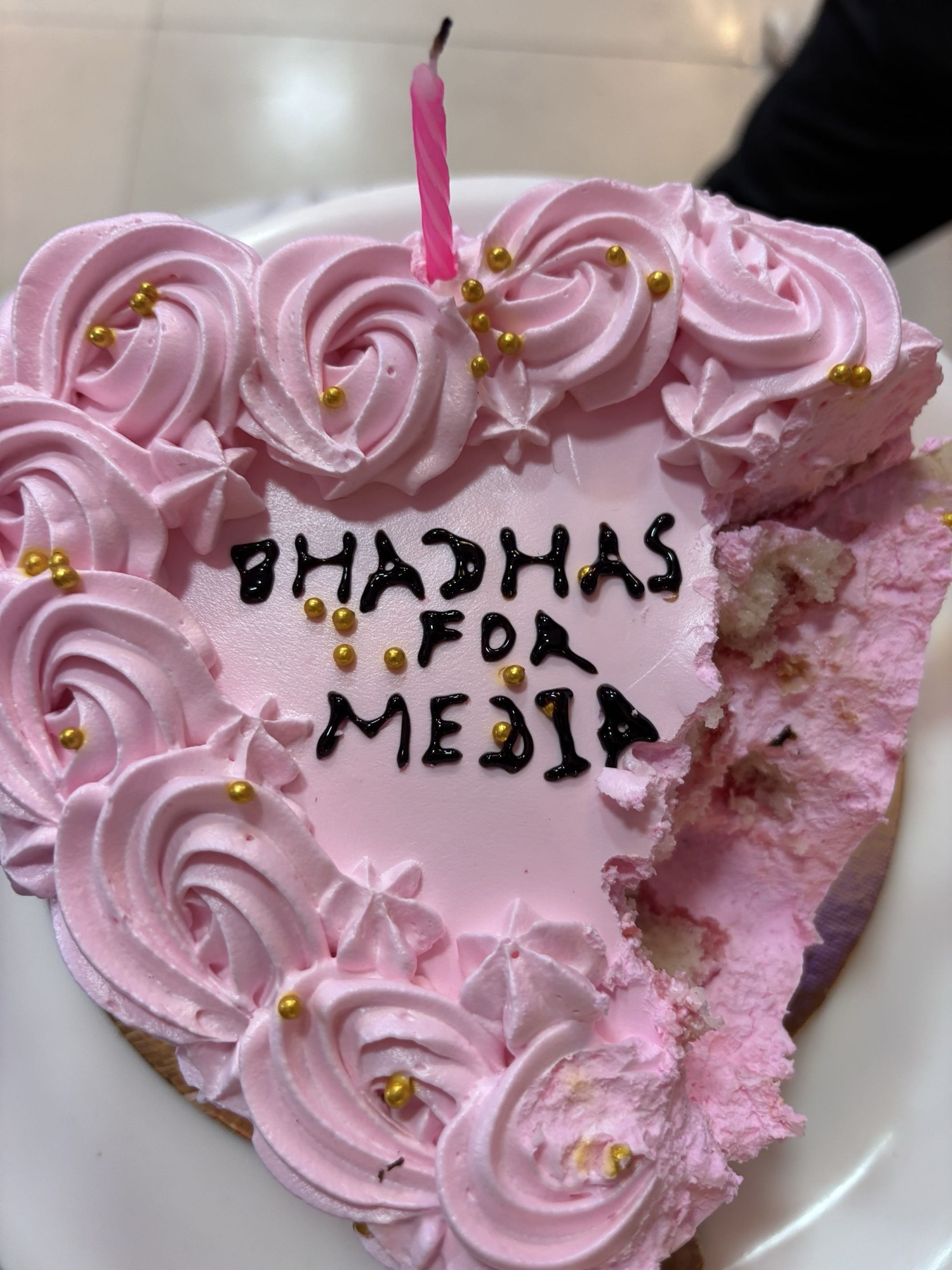 Pink frosted cake with many rose-like swirls and gold beads, a single pink candle, and a bite taken from the side; message partially visible on the cake surface.