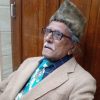 Older man with gray hair and mustache wearing glasses, a tan blazer, blue plaid shirt, turquoise patterned tie, and a fuzzy hat, seated by a wooden door.