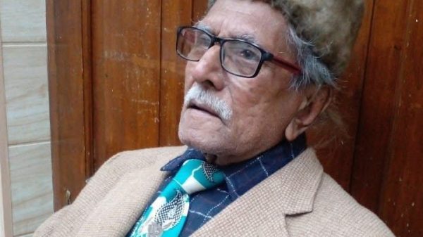 Older man with gray hair and mustache wearing glasses, a tan blazer, blue plaid shirt, turquoise patterned tie, and a fuzzy hat, seated by a wooden door.