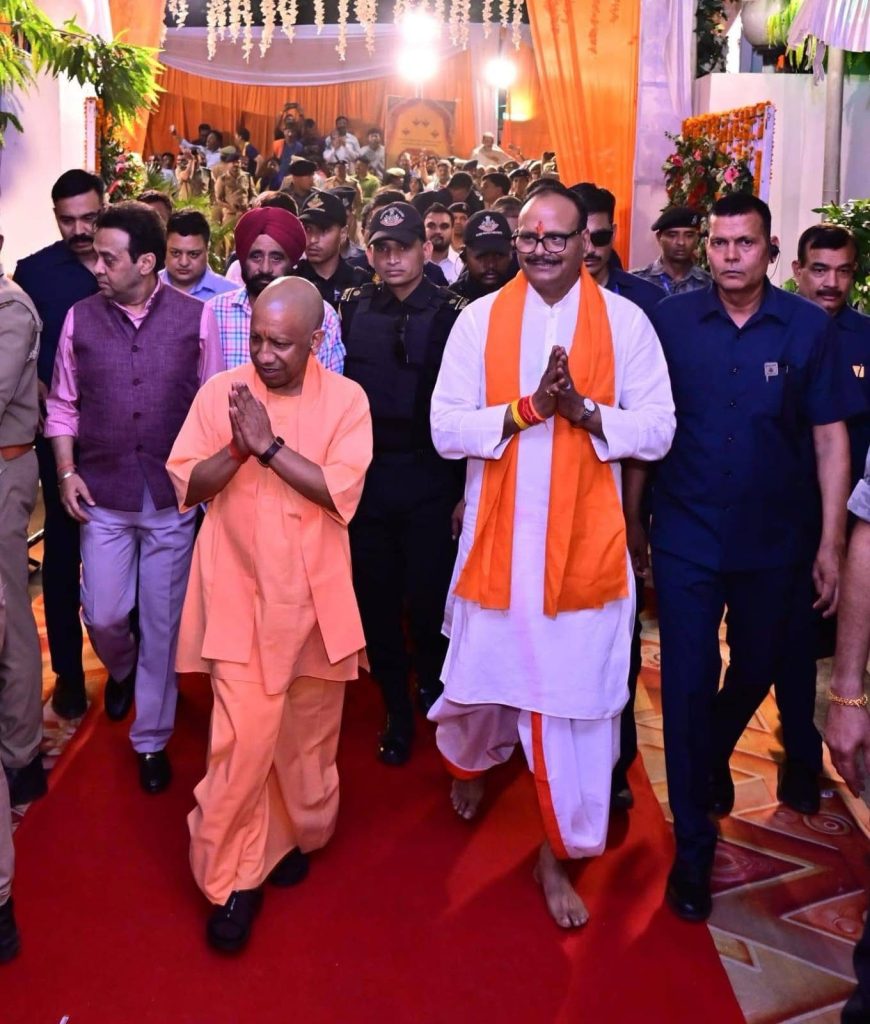Politician in white kurta with an orange scarf walks on a red carpet, hands in a greeting pose, flanked by security at an indoor ceremony.
