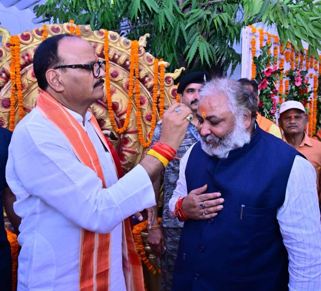 Man applying a ceremonial tilak to an older man at a festive event, with orange garlands and floral decor in the background.