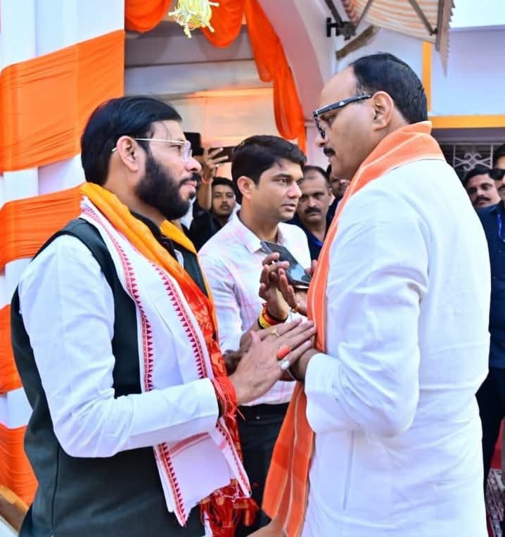 Two men wearing white shirts and orange scarves talk warmly at a festive event with orange drapes in the background.