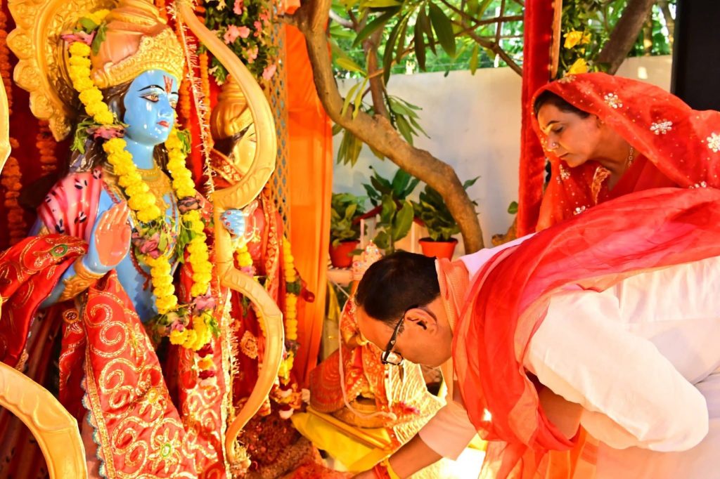 Blue Krishna idol decorated with yellow flower garlands; worshippers in orange and pink robes bow in reverence at a colorful temple altar.