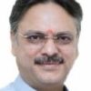 Head-and-shoulders portrait of a middle-aged Indian man with glasses and a mustache, wearing a light blue kurta and a red tilak on his forehead against a plain background.