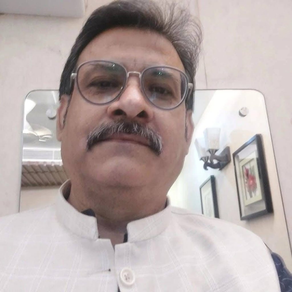Close-up portrait of a man with gray mustache and round glasses wearing a light traditional shirt, indoors in front of a mirror and wall art.