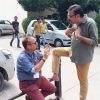 Two men in casual clothes converse outdoors; one kneels on a railing with hands gesturing, the other stands with a foot on the railing, nearby car visible.