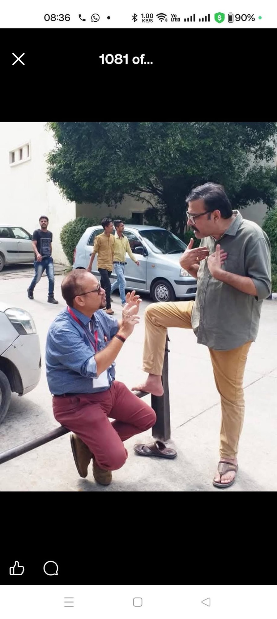 Two men in casual clothes converse outdoors; one kneels on a railing with hands gesturing, the other stands with a foot on the railing, nearby car visible.