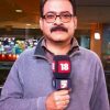 Male reporter holding a microphone with news channel logos in a newsroom setting; looking at camera with glasses and mustache