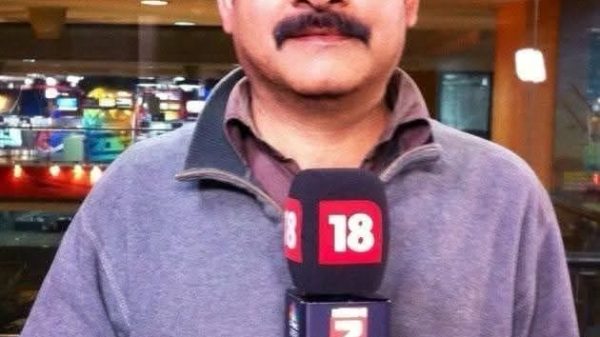 Male reporter holding a microphone with news channel logos in a newsroom setting; looking at camera with glasses and mustache