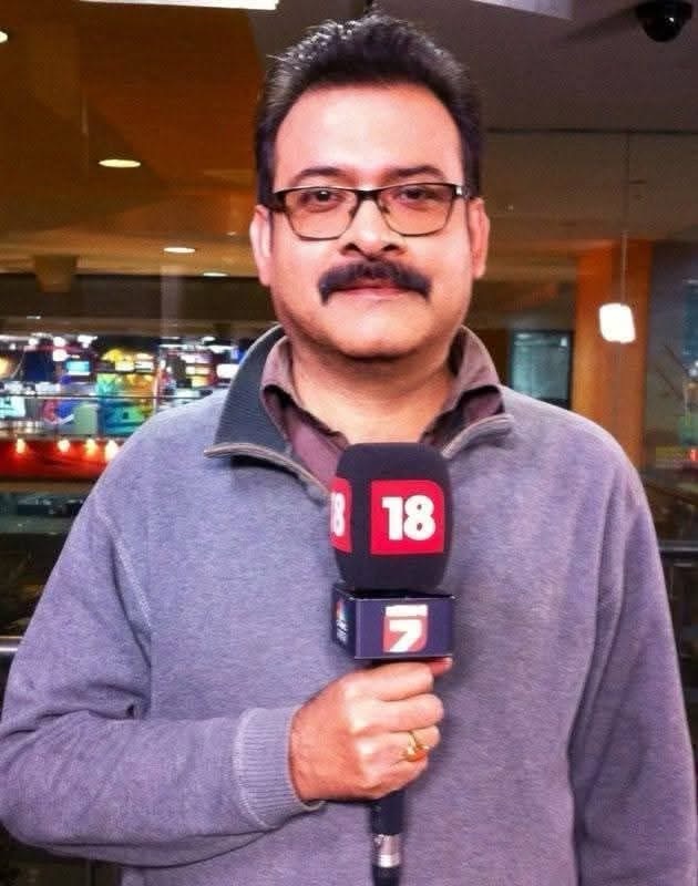 img_8483 | No. 1 Indian Media News Portal Male reporter holding a microphone with news channel logos in a newsroom setting; looking at camera with glasses and mustache