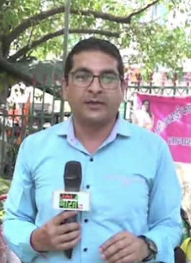 img_8568 | No. 1 Indian Media News Portal Male reporter in a light blue shirt holding a microphone with a news logo, standing outdoors in a sunny, leafy area.