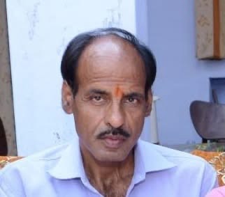 img_8574 | No. 1 Indian Media News Portal Portrait of a middle-aged man with an orange tilak on his forehead, wearing a light blue shirt, seated indoors with a pale wall in the background.