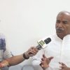 News interview scene: journalist with glasses interviews a man in a white kurta; microphone labeled with JAI BHARAT TV logo in the corner.