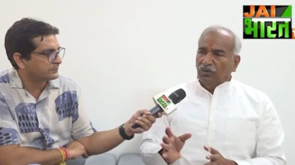 News interview scene: journalist with glasses interviews a man in a white kurta; microphone labeled with JAI BHARAT TV logo in the corner.