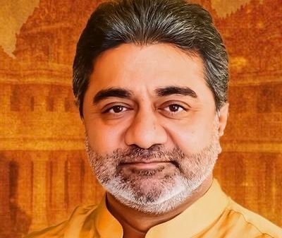 Portrait of a middle-aged man with a salt-and-pepper beard wearing a saffron kurta, smiling, against an warm orange-toned background resembling a temple setting.