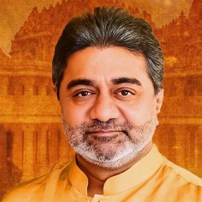 Portrait of a middle-aged man with a salt-and-pepper beard wearing a saffron kurta, smiling, against an warm orange-toned background resembling a temple setting.
