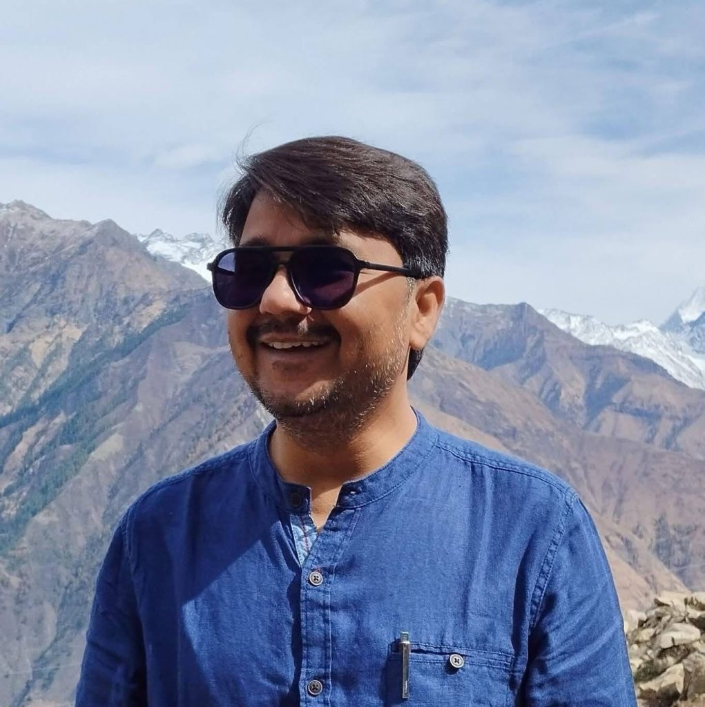 img_8600 | No. 1 Indian Media News Portal Smiling man wearing dark sunglasses and a blue shirt with snow-capped mountains behind him.