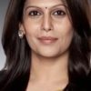 Portrait of a smiling Indian woman with dark hair, wearing a gold and beige saree and a small bindi against a neutral background.