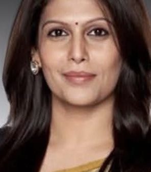 Portrait of a smiling Indian woman with dark hair, wearing a gold and beige saree and a small bindi against a neutral background.