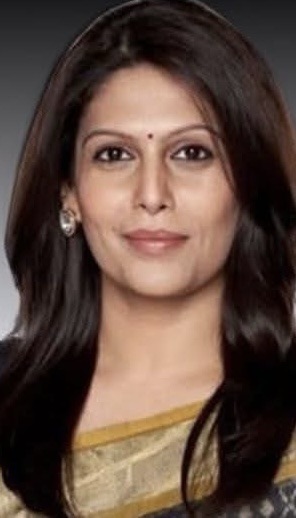 Portrait of a smiling Indian woman with dark hair, wearing a gold and beige saree and a small bindi against a neutral background.
