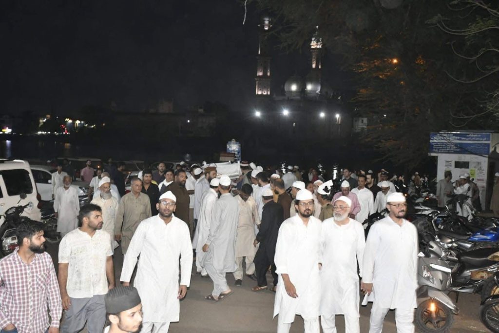 Crowd of men in white traditional clothing gathering outdoors at night near a river with a illuminated mosque in the background.