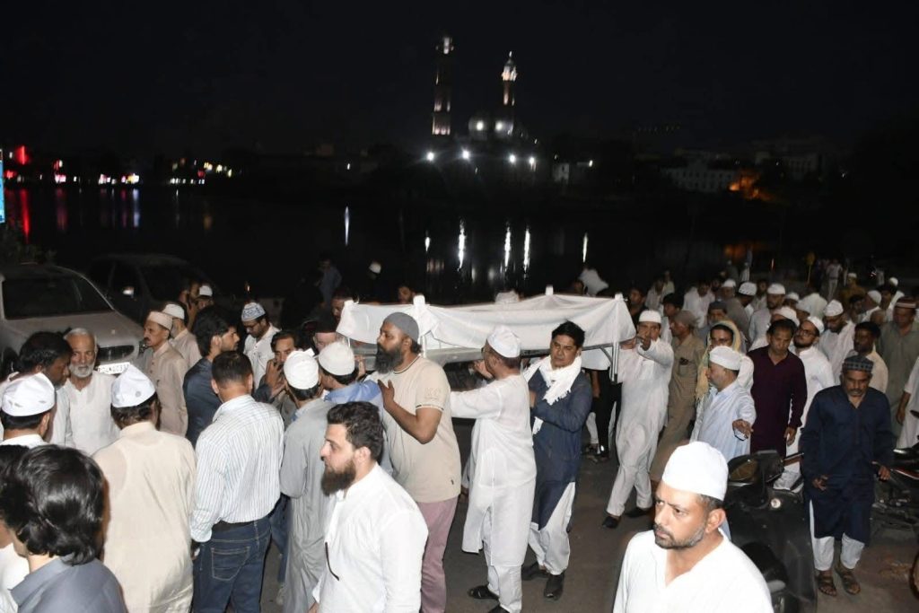 Nighttime funeral procession with many men in white garments carrying a white coffin along a riverside street.