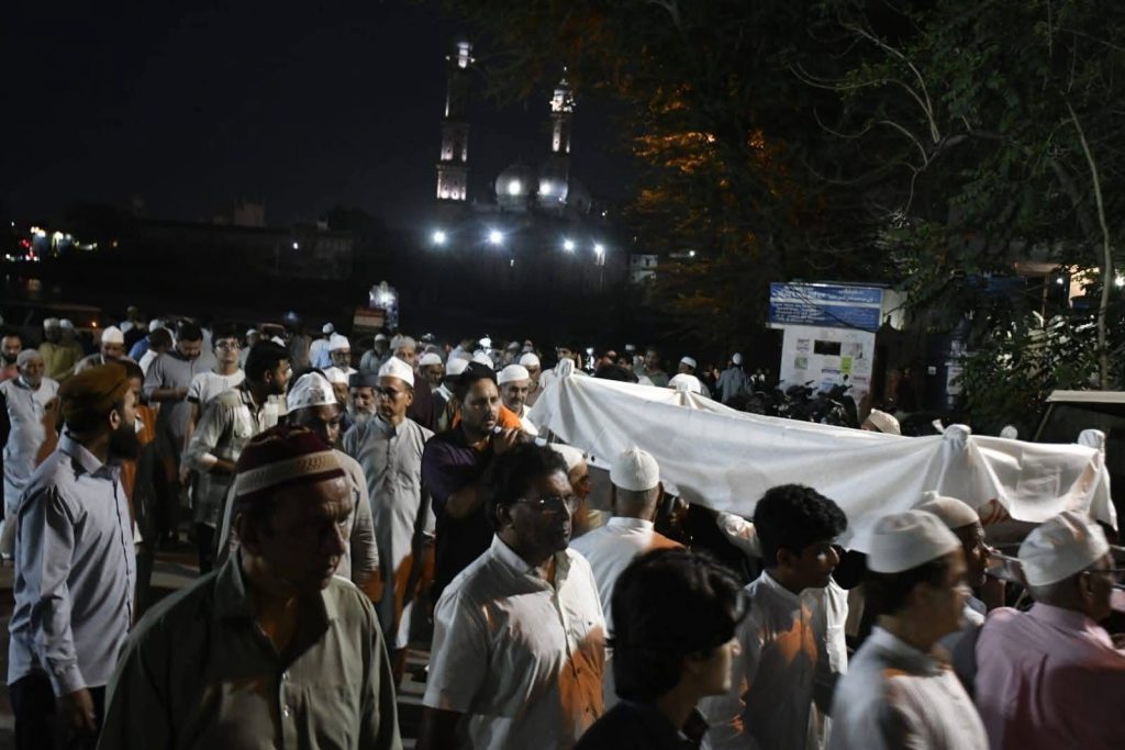 Nighttime funeral procession with men carrying a white-shrouded coffin through a crowd; a lit mosque in the background.