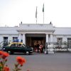 White colonial-style government building with flagpoles, a vintage green-and-yellow taxi parked in front, and people near the entrance.