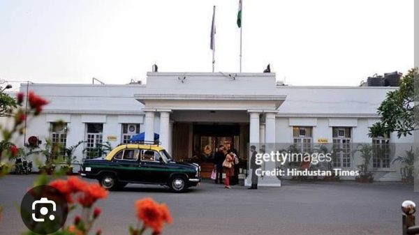 White colonial-style government building with flagpoles, a vintage green-and-yellow taxi parked in front, and people near the entrance.