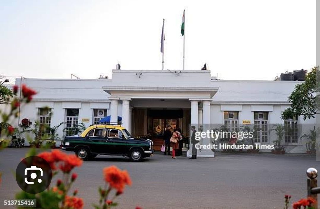 White colonial-style government building with flagpoles, a vintage green-and-yellow taxi parked in front, and people near the entrance.