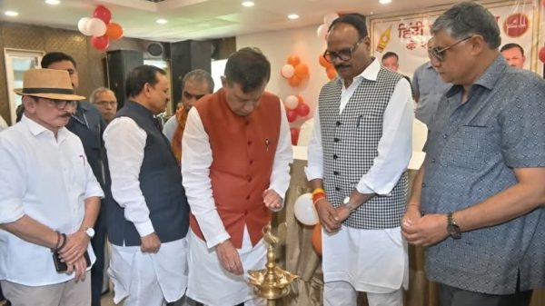 Group of men in traditional Indian attire lighting a ceremonial brass lamp at an indoor event, with balloons and a banner in the background.