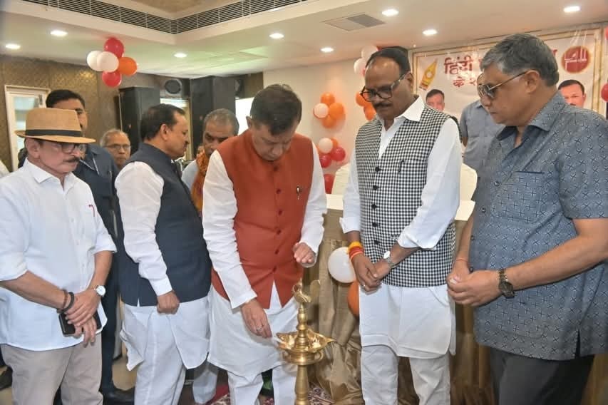 Group of men in traditional Indian attire lighting a ceremonial brass lamp at an indoor event, with balloons and a banner in the background.