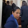 Woman in blue blazer with glasses looking toward camera in a hallway with people in business attire in the background, wearing a lanyard.