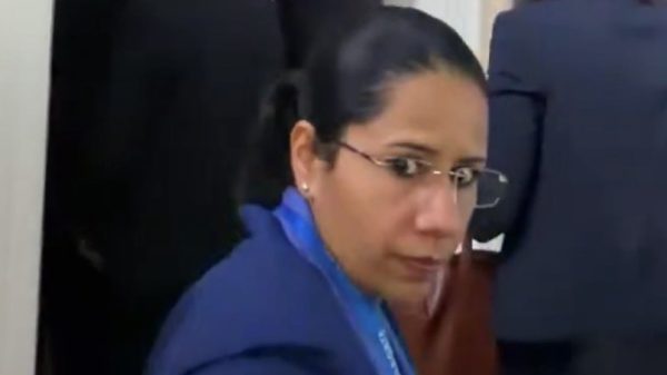 Woman in blue blazer with glasses looking toward camera in a hallway with people in business attire in the background, wearing a lanyard.