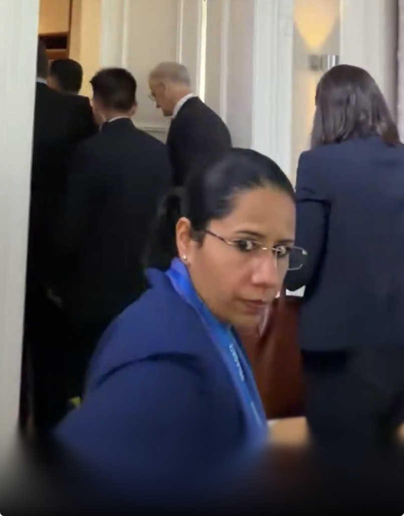 Woman in blue blazer with glasses looking toward camera in a hallway with people in business attire in the background, wearing a lanyard.