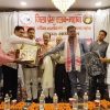 Group of men on a stage exchanging awards during a formal ceremony, with a banner in Hindi in the background and marigold decorations on the table in front.
