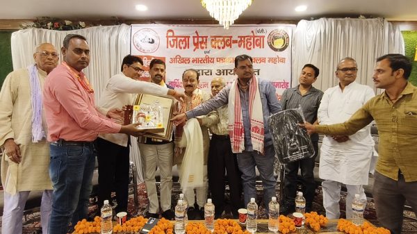 Group of men on a stage exchanging awards during a formal ceremony, with a banner in Hindi in the background and marigold decorations on the table in front.