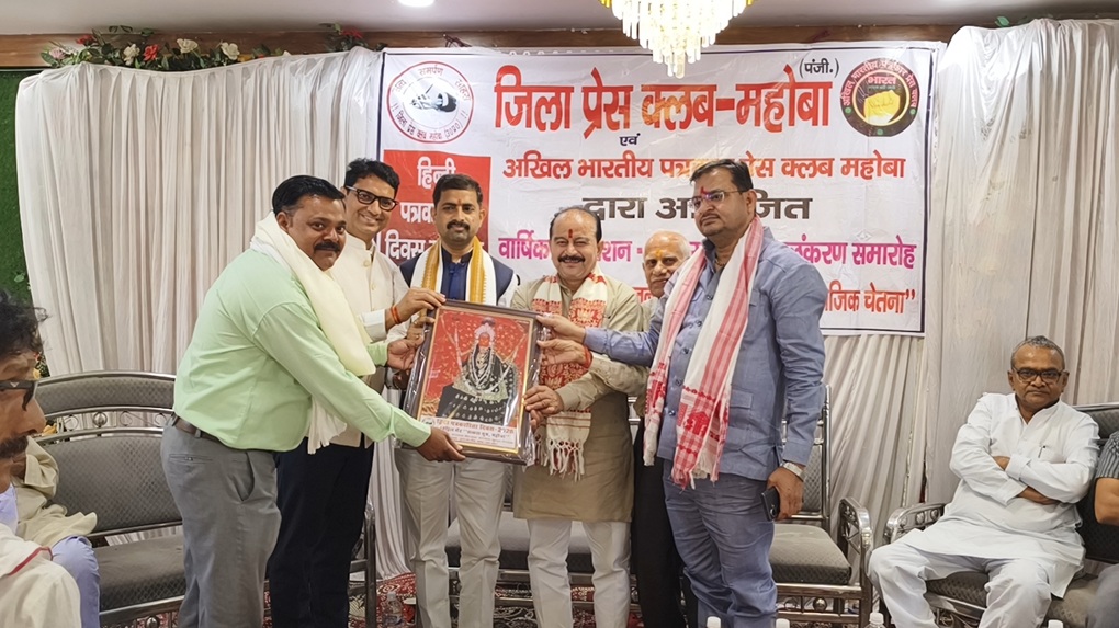 mahoba_ | No. 1 Indian Media News Portal Group of men at a formal ceremony presenting a framed portrait to a recipient, with a Hindi banner and seated guests in the background.