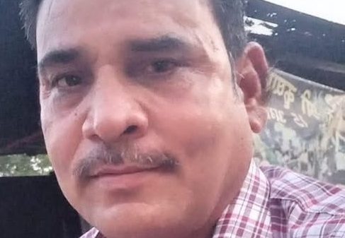 Man with short dark hair and mustache wearing a red and white plaid shirt, posing for a selfie outdoors.