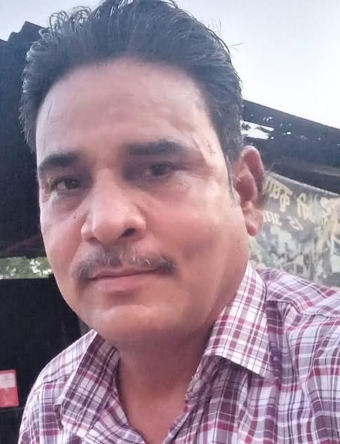 Man with short dark hair and mustache wearing a red and white plaid shirt, posing for a selfie outdoors.