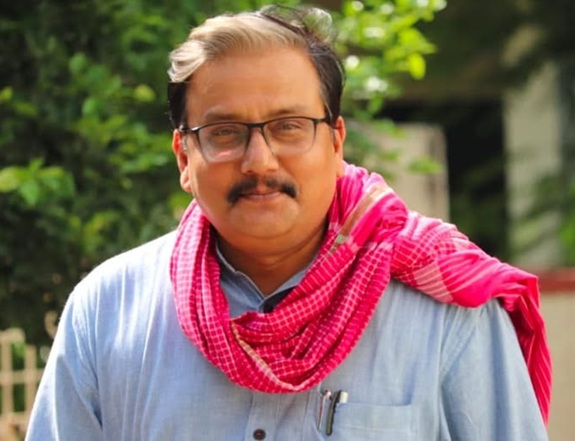 manoj_jha | No. 1 Indian Media News Portal Man with glasses and mustache wearing a light blue shirt and pink scarf, outdoors with greenery in the background.