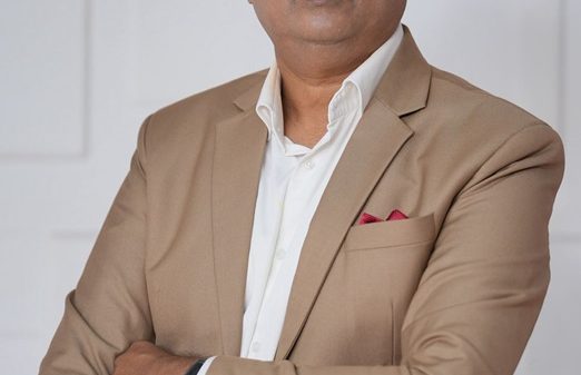 Professional portrait of a middle-aged man with gray hair, wearing a tan suit and white shirt, arms crossed against a light, neutral background.